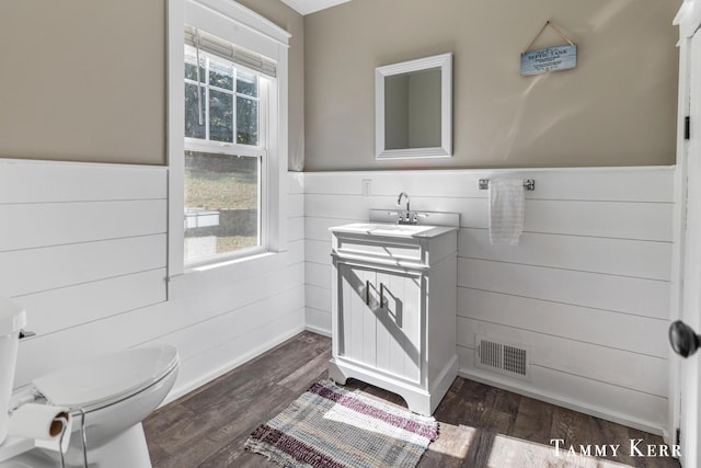 half bath with visible vents, toilet, wood finished floors, wainscoting, and vanity