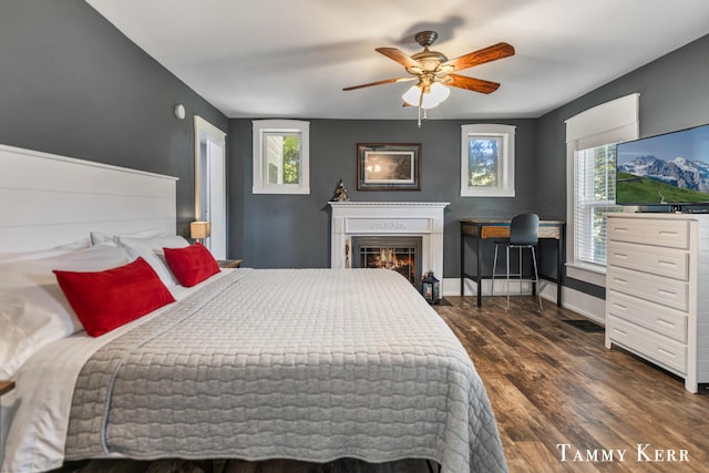 bedroom with ceiling fan, baseboards, dark wood-type flooring, and a glass covered fireplace