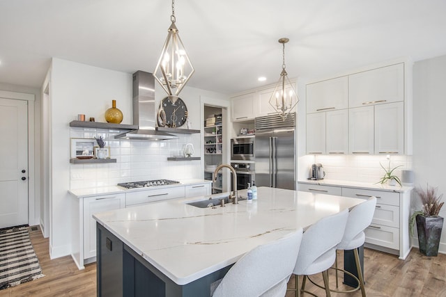 kitchen with open shelves, wall chimney range hood, built in appliances, white cabinetry, and a sink