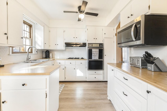 kitchen with a sink, stainless steel microwave, under cabinet range hood, backsplash, and light wood finished floors