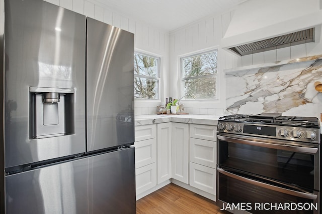 kitchen featuring light wood finished floors, custom range hood, white cabinets, backsplash, and appliances with stainless steel finishes