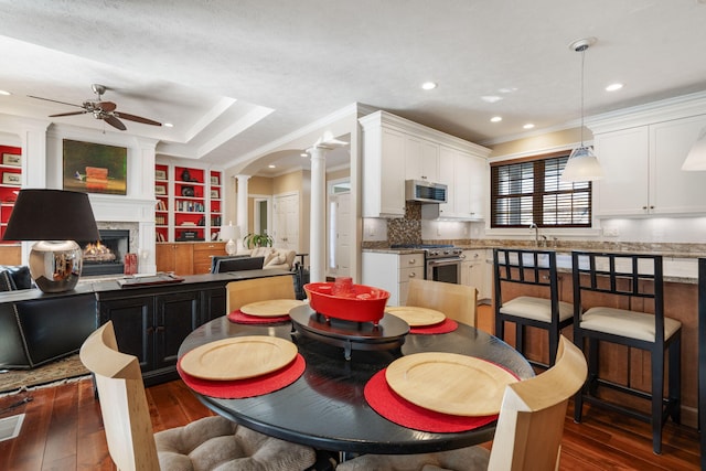 dining space with decorative columns, a lit fireplace, dark wood finished floors, ceiling fan, and ornamental molding