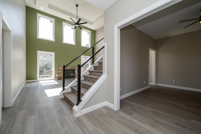 staircase featuring a towering ceiling, a raised ceiling, wood finished floors, a ceiling fan, and baseboards