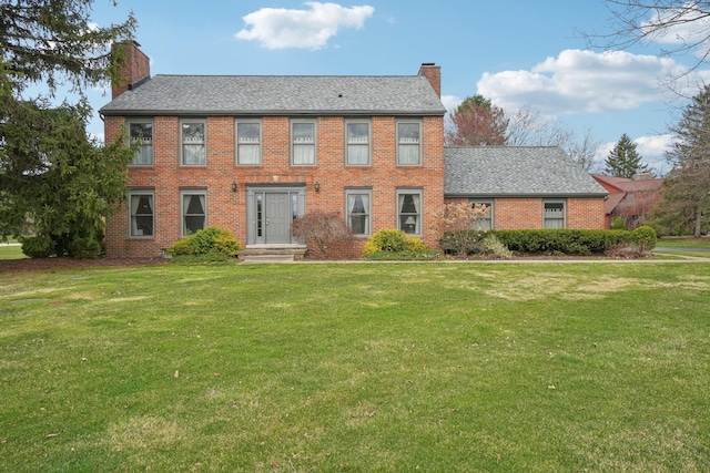 colonial house featuring roof with shingles, a chimney, a front yard, and brick siding