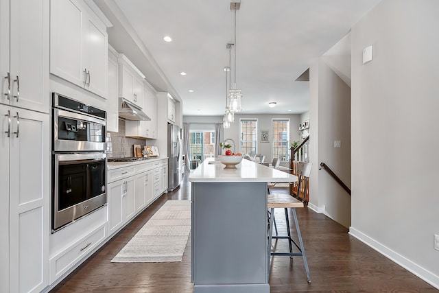 kitchen with an island with sink, appliances with stainless steel finishes, white cabinetry, baseboards, and under cabinet range hood