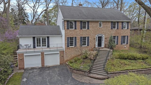 colonial-style house featuring brick siding, a chimney, roof with shingles, and aphalt driveway