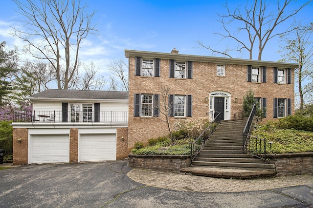 colonial-style house featuring brick siding, a garage, a chimney, and aphalt driveway