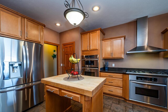 kitchen with wood counters, appliances with stainless steel finishes, stone tile flooring, a center island, and wall chimney exhaust hood