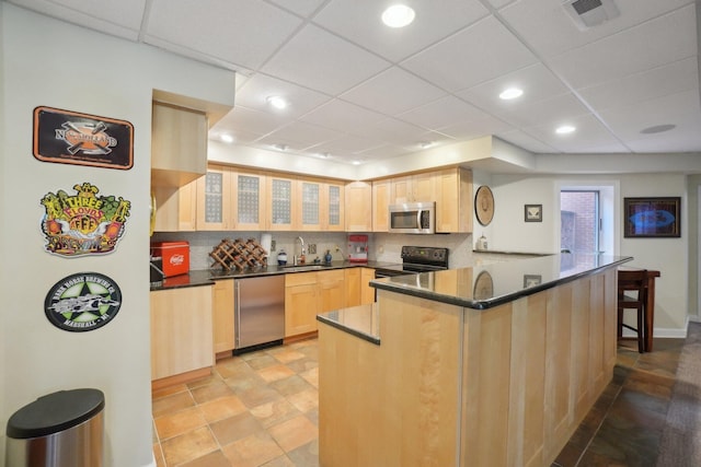 kitchen with dark countertops, a sink, light brown cabinetry, stainless steel appliances, and a peninsula