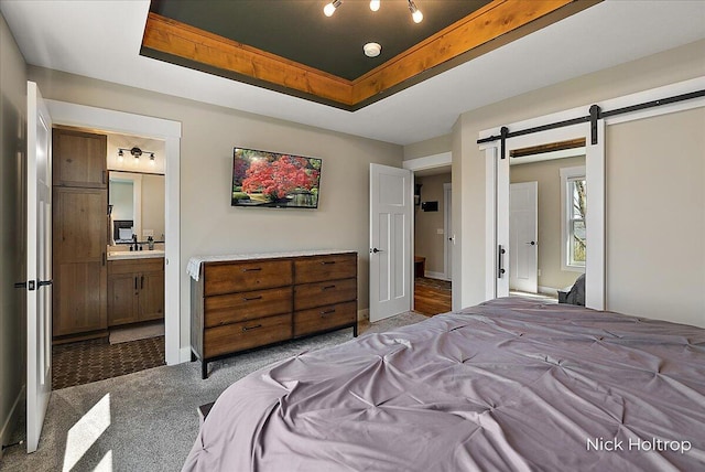 bedroom with a barn door, a tray ceiling, dark carpet, connected bathroom, and a sink