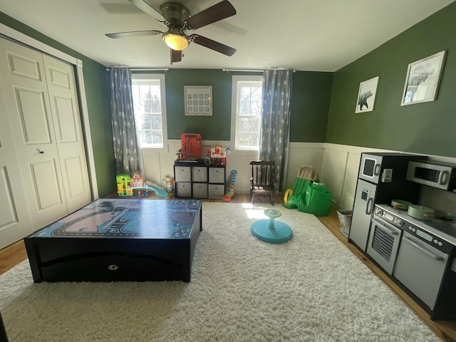 recreation room featuring wood finished floors, ceiling fan, and wainscoting