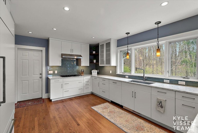 kitchen with pendant lighting, wood finished floors, a sink, black cooktop, and under cabinet range hood