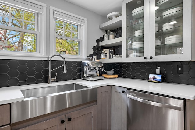 kitchen with backsplash, dishwasher, a sink, light countertops, and glass insert cabinets