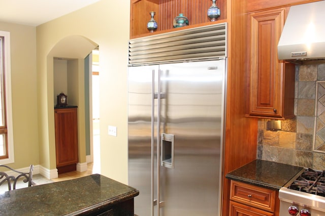 60 Timber Ridge Ct, Milan, MI 48160 - kitchen featuring built in fridge, wall chimney range hood, arched walkways, backsplash, and dark stone countertops