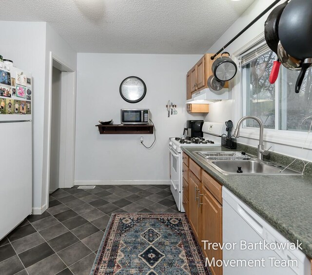 kitchen with dark tile patterned floors, brown cabinets, a sink, under cabinet range hood, and white appliances