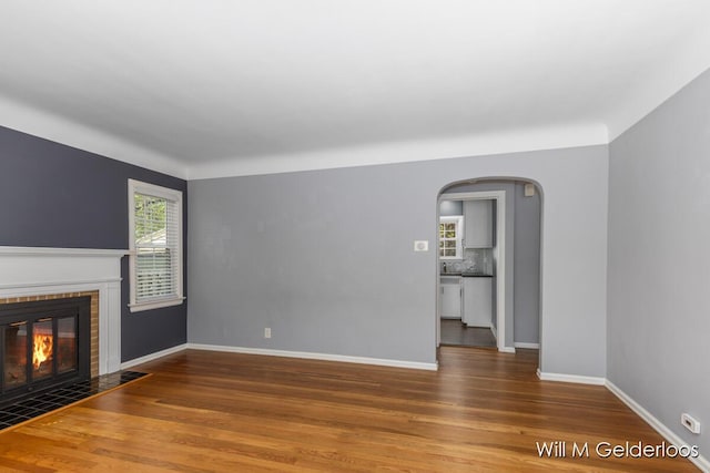 unfurnished living room with arched walkways, a fireplace with flush hearth, wood finished floors, and baseboards