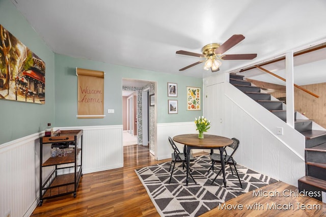 dining room with wood finished floors, a ceiling fan, wainscoting, and stairs