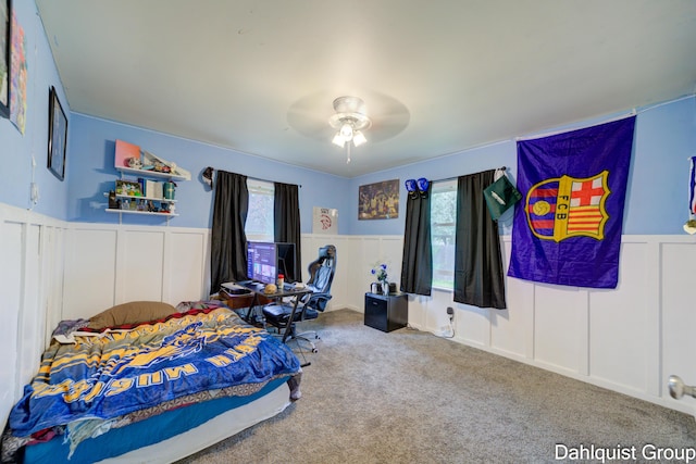 carpeted bedroom featuring wainscoting, a decorative wall, multiple windows, and ceiling fan