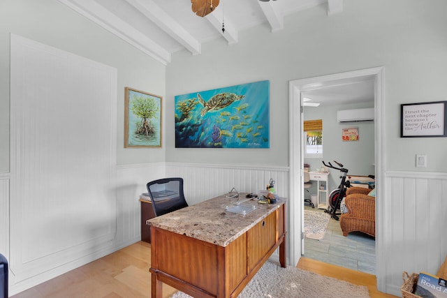 office area with a wall mounted air conditioner, beam ceiling, light wood-type flooring, and a wainscoted wall