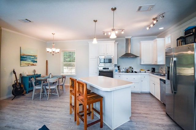 kitchen with wall chimney range hood, appliances with stainless steel finishes, white cabinetry, a kitchen island, and decorative light fixtures
