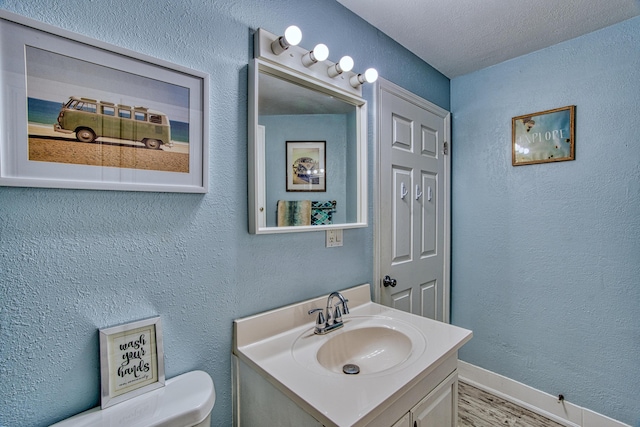 half bath with vanity, toilet, a textured wall, and baseboards