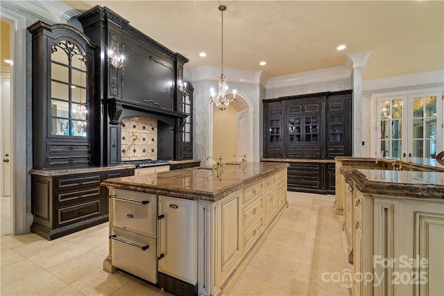 kitchen featuring ornamental molding, cream cabinets, an island with sink, and dark stone countertops