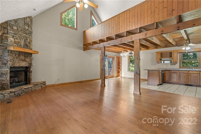 unfurnished living room with ceiling fan with notable chandelier, beamed ceiling, high vaulted ceiling, light hardwood / wood-style flooring, and a stone fireplace