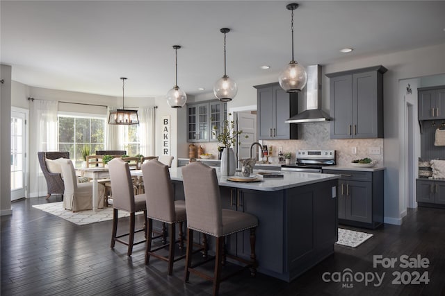 kitchen featuring stainless steel range with electric stovetop, gray cabinetry, a kitchen island with sink, wall chimney range hood, and decorative light fixtures