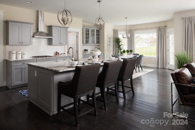 kitchen featuring gray cabinetry, a center island with sink, a kitchen breakfast bar, wall chimney range hood, and stainless steel gas cooktop