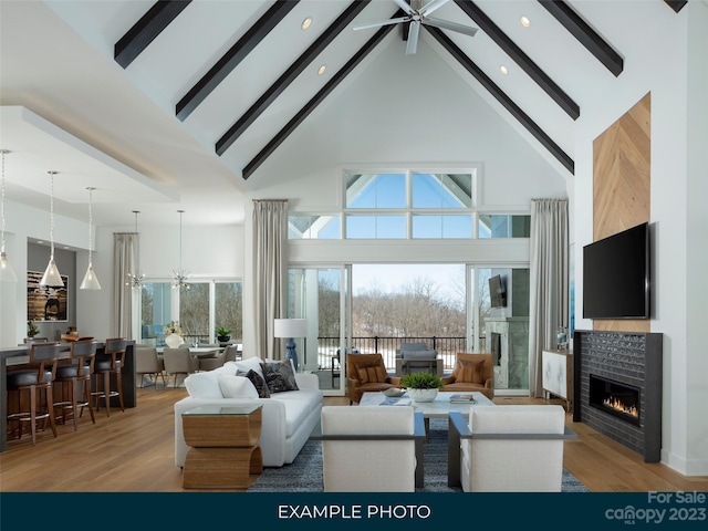 living room with light wood-type flooring, beam ceiling, a brick fireplace, ceiling fan with notable chandelier, and high vaulted ceiling