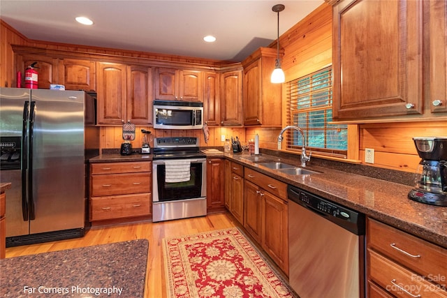 kitchen featuring sink, dark stone counters, stainless steel appliances, decorative light fixtures, and light wood-type flooring