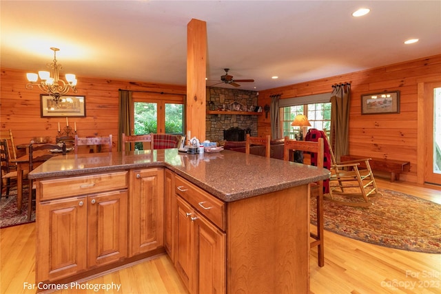 kitchen featuring wood walls, ceiling fan with notable chandelier, light wood-type flooring, and a stone fireplace