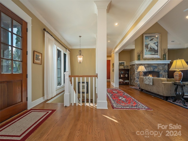 foyer entrance featuring decorative columns, light hardwood / wood-style floors, a chandelier, a stone fireplace, and crown molding