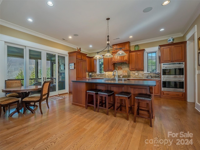 kitchen with hanging light fixtures, backsplash, double oven, a kitchen island with sink, and light hardwood / wood-style floors