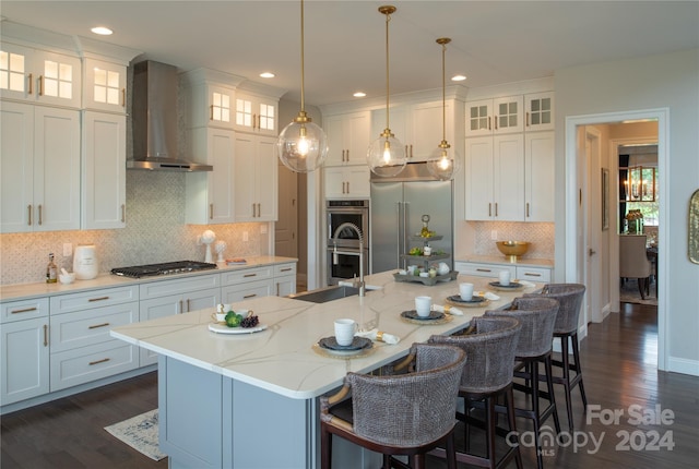 kitchen featuring wall chimney range hood, decorative light fixtures, an island with sink, appliances with stainless steel finishes, and white cabinetry