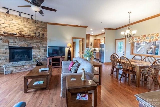 living room with rail lighting, ornamental molding, wood-type flooring, ceiling fan with notable chandelier, and a stone fireplace