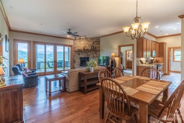 dining area featuring a stone fireplace, crown molding, a mountain view, light wood-type flooring, and ceiling fan with notable chandelier