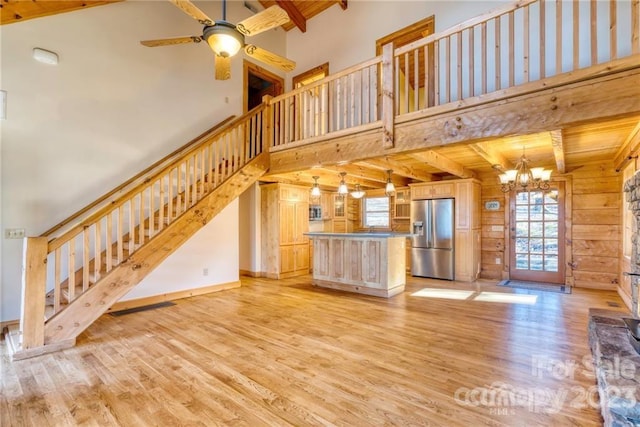 unfurnished living room featuring a towering ceiling, light wood-type flooring, wooden walls, ceiling fan with notable chandelier, and beam ceiling