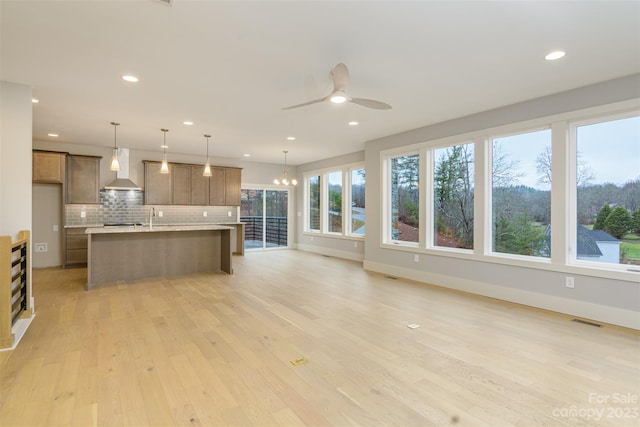 kitchen with a center island with sink, light wood-type flooring, pendant lighting, wall chimney range hood, and backsplash