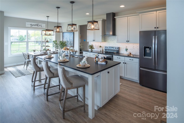 kitchen featuring wall chimney exhaust hood, stainless steel appliances, a kitchen island with sink, decorative light fixtures, and white cabinetry