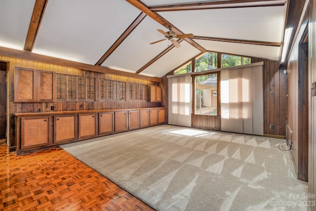 unfurnished living room with high vaulted ceiling, ceiling fan, beamed ceiling, wooden walls, and light carpet