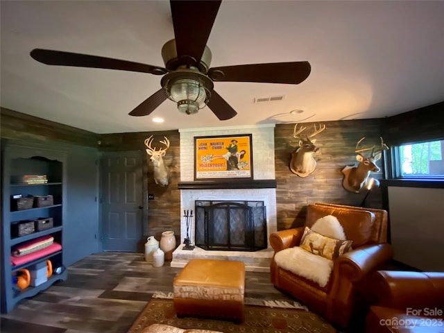 sitting room with ceiling fan, wooden walls, a stone fireplace, and dark hardwood / wood-style floors