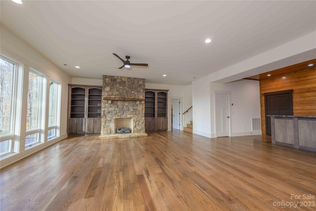 unfurnished living room featuring ceiling fan, a wealth of natural light, built in shelves, a stone fireplace, and hardwood / wood-style flooring