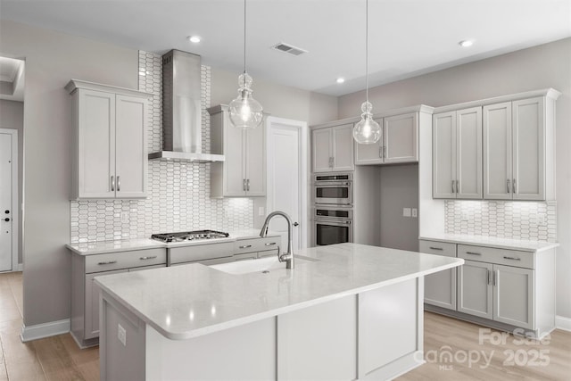 kitchen with decorative light fixtures, a center island with sink, appliances with stainless steel finishes, a sink, and wall chimney exhaust hood