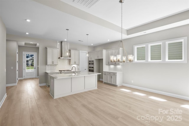 kitchen featuring visible vents, an island with sink, wall chimney exhaust hood, light countertops, and pendant lighting