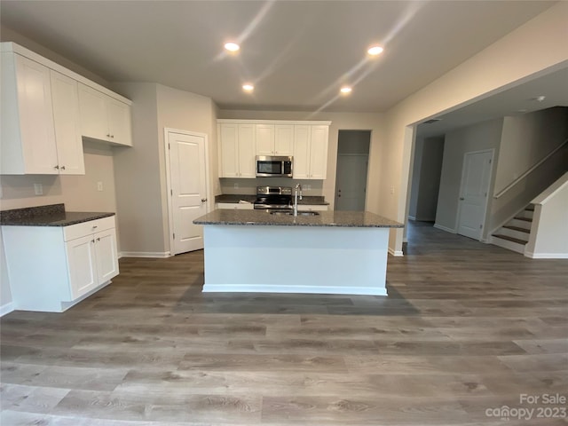 kitchen with appliances with stainless steel finishes, dark stone counters, an island with sink, wood-type flooring, and white cabinets