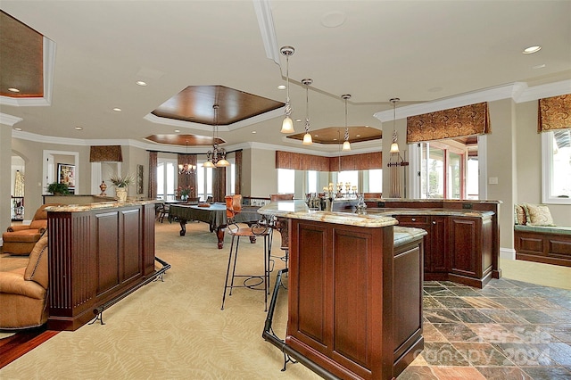 kitchen with a breakfast bar area, hanging light fixtures, a raised ceiling, a kitchen island with sink, and light stone counters