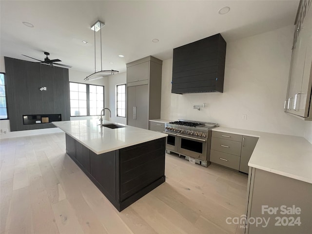 kitchen with ventilation hood, a kitchen island with sink, sink, light hardwood / wood-style flooring, and range with two ovens