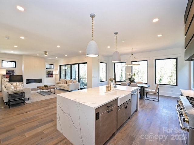 kitchen with sink, light stone counters, wood-type flooring, hanging light fixtures, and an island with sink