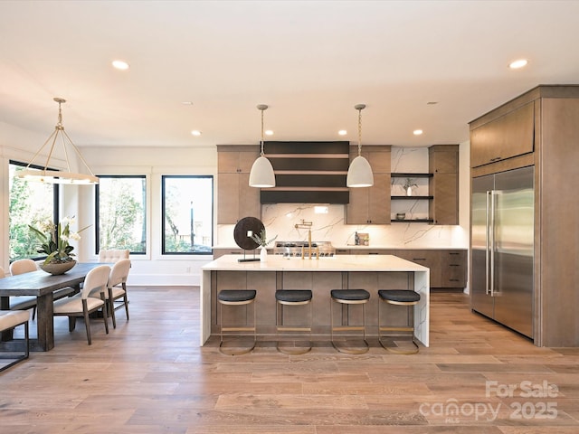 kitchen with a center island with sink, light hardwood / wood-style flooring, stainless steel built in fridge, pendant lighting, and backsplash